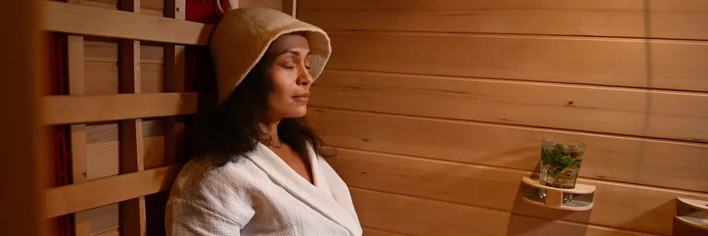 A woman in a robe and a felt hat sitting with her eyes closed inside a wood-lined infrared cabin.