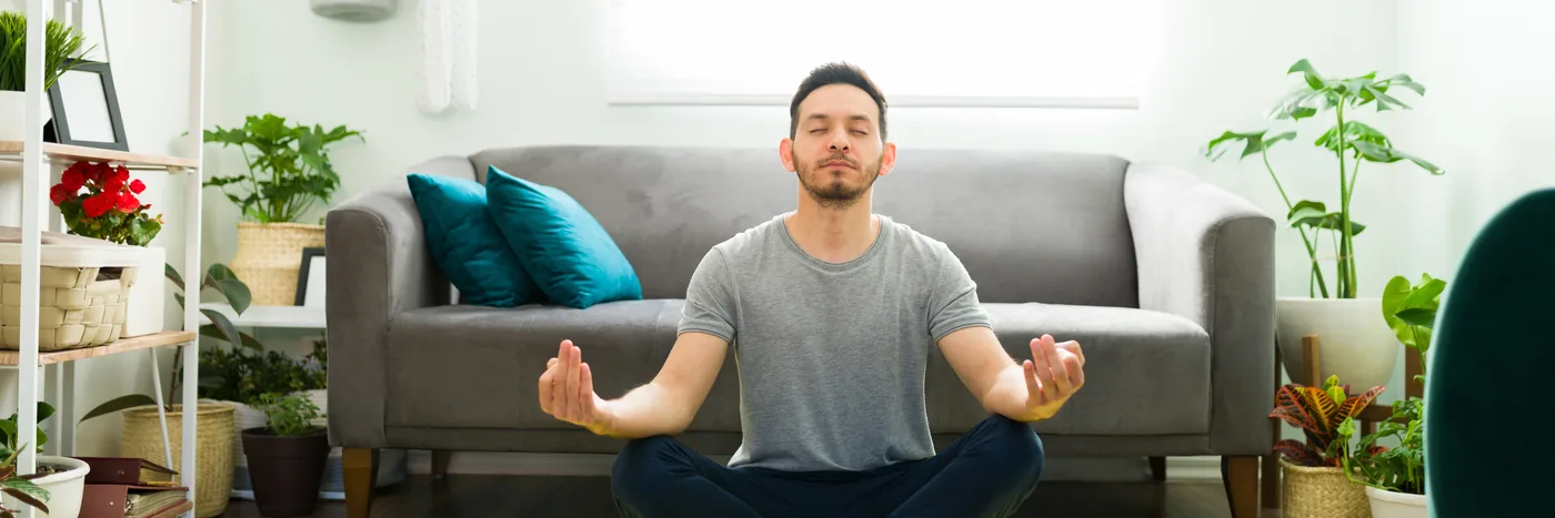 A man sitting cross-legged and upright on a mat in his living room with his eyes closed.